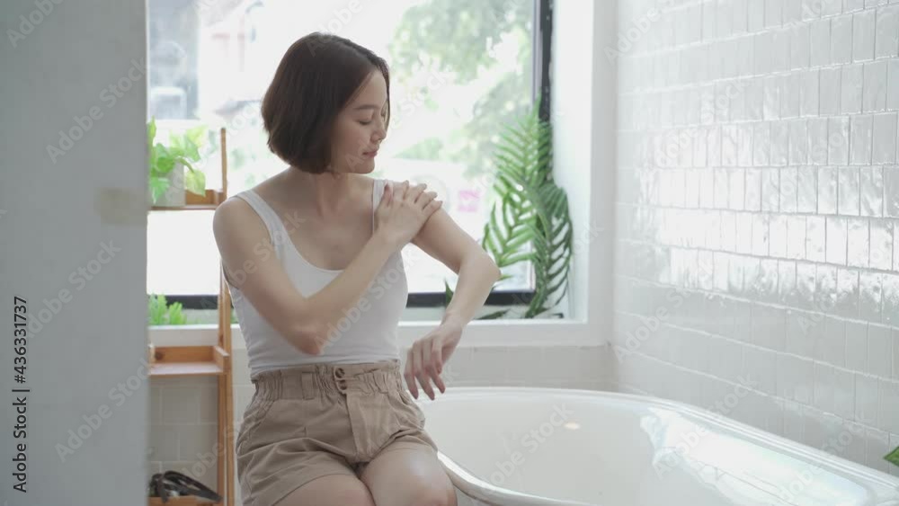 Asian woman short hair, applying cream lotion on hands, leg, and arm in white bathroom near bathtub at home studio decor some green plant. white cream rubbing to treatment skin condition. scrubbing Asian woman short hair, applying cream lotion on hands, leg, and arm in white bathroom near bathtub at home studio decor some green plant. white cream rubbing to treatment skin condition. scrubbing