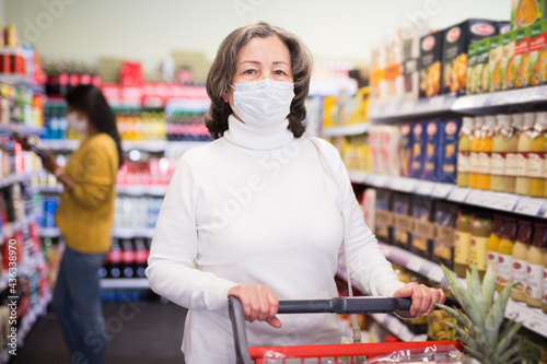 Woman in protective mask with shopping cart choosing groceries in supermarket