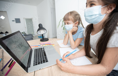Cute little girl wearing a medical mask on her face doing distance learning using a laptop at home in the bedroom. A girl in a protective mask on her face holds her head in her hand. Distance Learning
