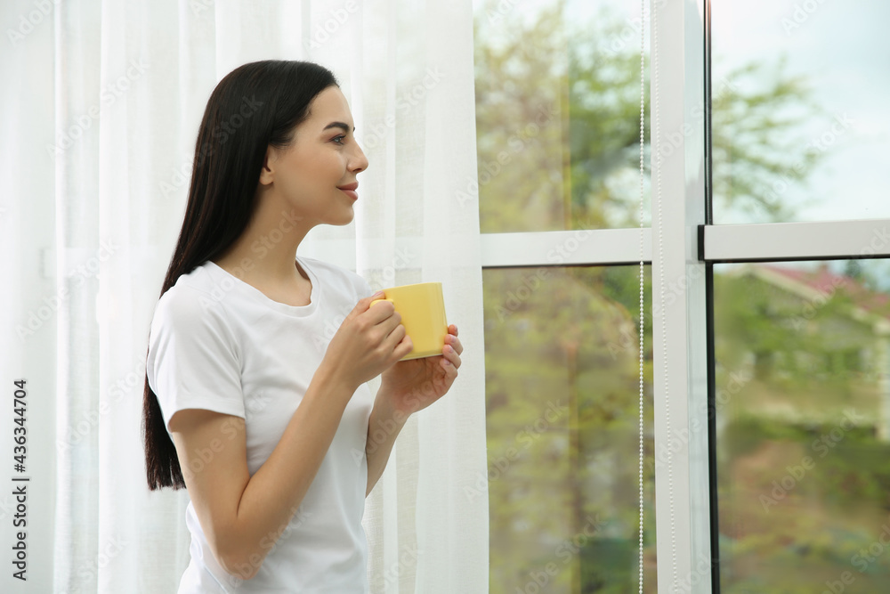 Young woman with cup of drink relaxing near window home, space for text Young woman with cup of drink relaxing near window home, space for text