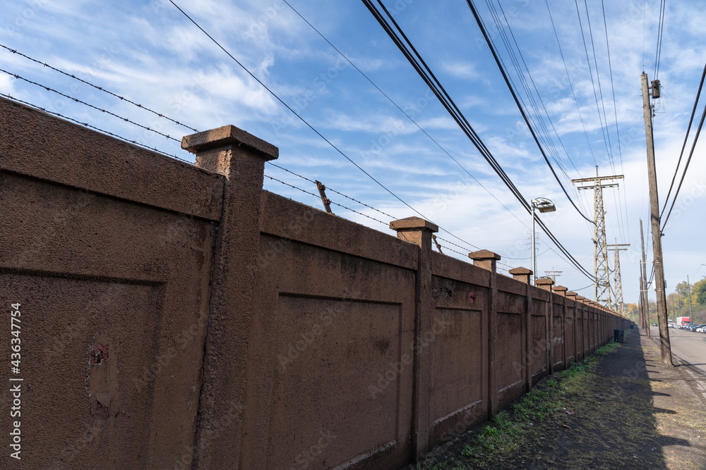Long view down a sidewalk bordered by a street and a tall stucco wall topped with barbed wire, blue sky with clouds industrial landscape, horizontal aspect