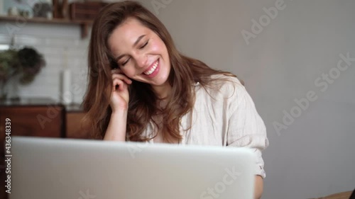 Young pretty woman talking on video call on laptop and waving hand while sitting at table inthe kitchen.home office, working remotely, freelancer, discussing a new project with team.