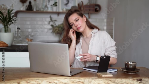 Young beautiful woman talking on a video call and waving her hand, putting on wireless headphones while sitting at the table in the kitchen.