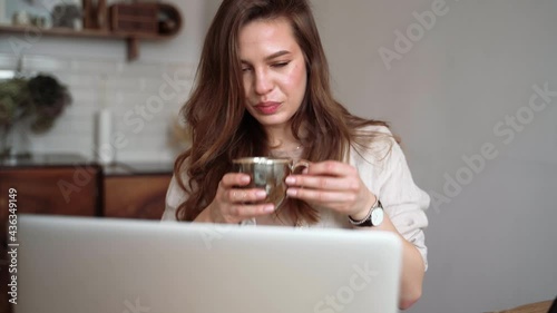 Smiling young blonde woman in beige linen clothes with her laptop computer, drinking coffee, sitting at a table indoors, talking on a video call. Homeschooling. Freelancer works from home