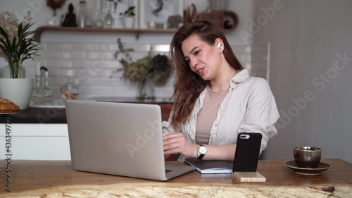 Young pretty woman talking on video call on laptop and waving hand while sitting at table inthe kitchen.home office, working remotely, freelancer, discussing a new project with team.