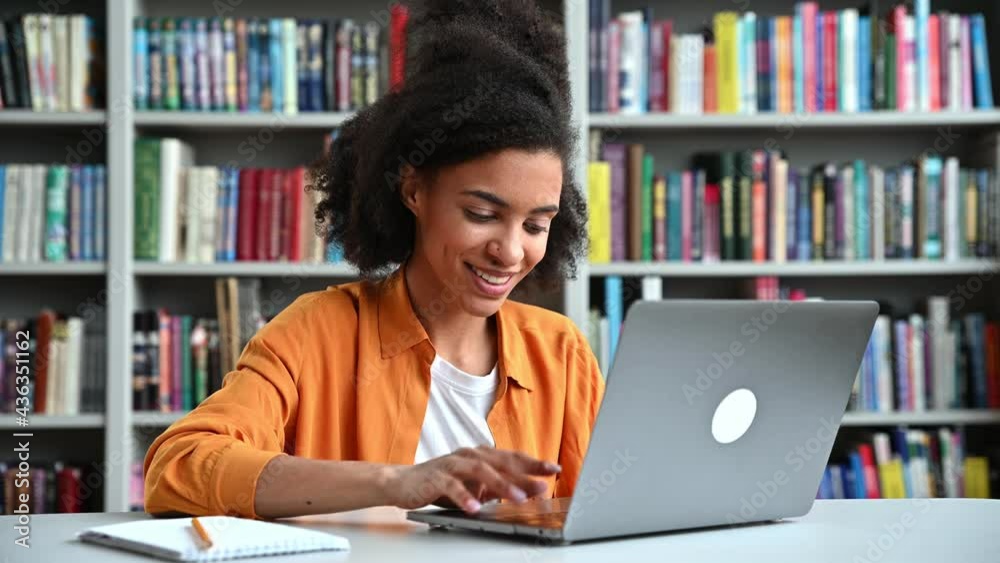 Happy african american smart female student with curly hair, trendy dressed, is sitting at a desk in the university library, uses laptop, typing on keyboard, focused taking notes during online lecture Happy african american smart female student with curly hair, trendy dressed, is sitting at a desk in the university library, uses laptop, typing on keyboard, focused taking notes during online lecture