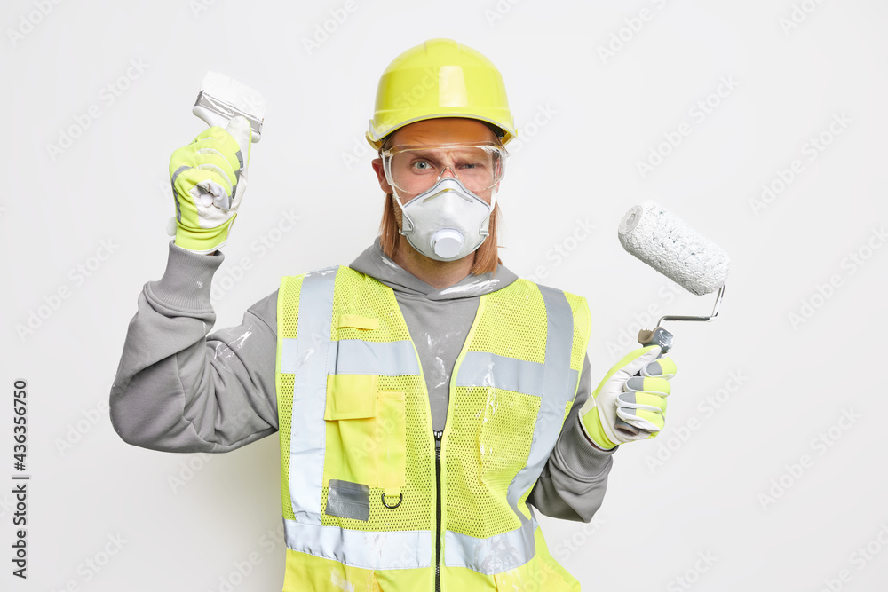 Construction and repair concept. Serious male builder uses painting tools busy doing house improvement wears protective respirator transparent glasses and uniform isolated over white background Construction and repair concept. Serious male builder uses painting tools busy doing house improvement wears protective respirator transparent glasses and uniform isolated over white background