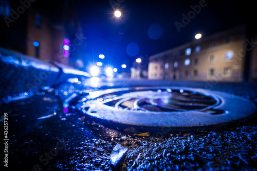 Night city after rain, the glowing lights of approaching car. Wide angle view of the level of a manhole on the pavement