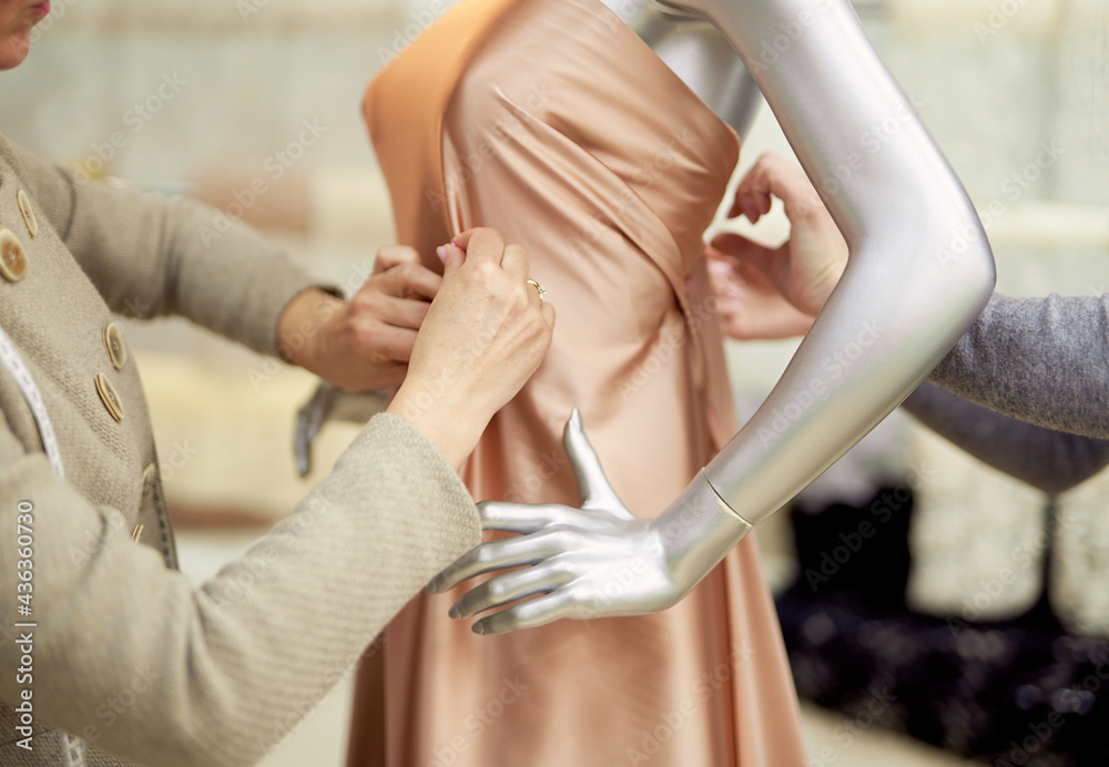 Fashion, dressmaking or tailoring concept. Professional team of tailors, dressmakers or designers working with new model applying linen fabrics beige cloth on mannequin or dummy in atelier