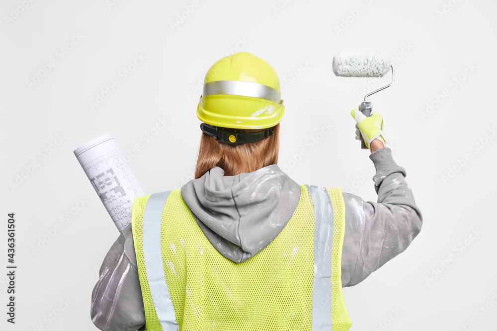 Unrecognizable man worker redecorates walls of new apartment holds roller and paper blueprint wears protective helmet and safety uniform stands back to camera isolated over white background. Unrecognizable man worker redecorates walls of new apartment holds roller and paper blueprint wears protective helmet and safety uniform stands back to camera isolated over white background.