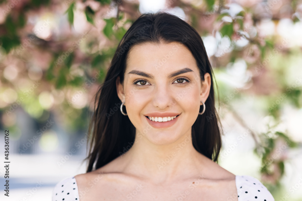 Portrait of a Gorgeous Dark Haired Woman Smiling Wearing Sammer Dress Charmingly while Standing at park on a background of sakura trees. Happy Young Woman Enjoys Life. Portrait of a Gorgeous Dark Haired Woman Smiling Wearing Sammer Dress Charmingly while Standing at park on a background of sakura trees. Happy Young Woman Enjoys Life.