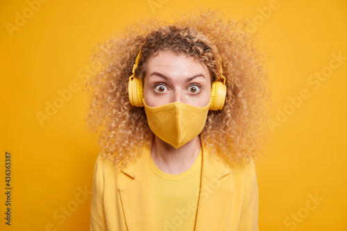 Surprised curly woman focused at camera wears protective face mask against coronavirus in public places listens music uses wireless headphones enjoys good sound quality isolated on yellow wall