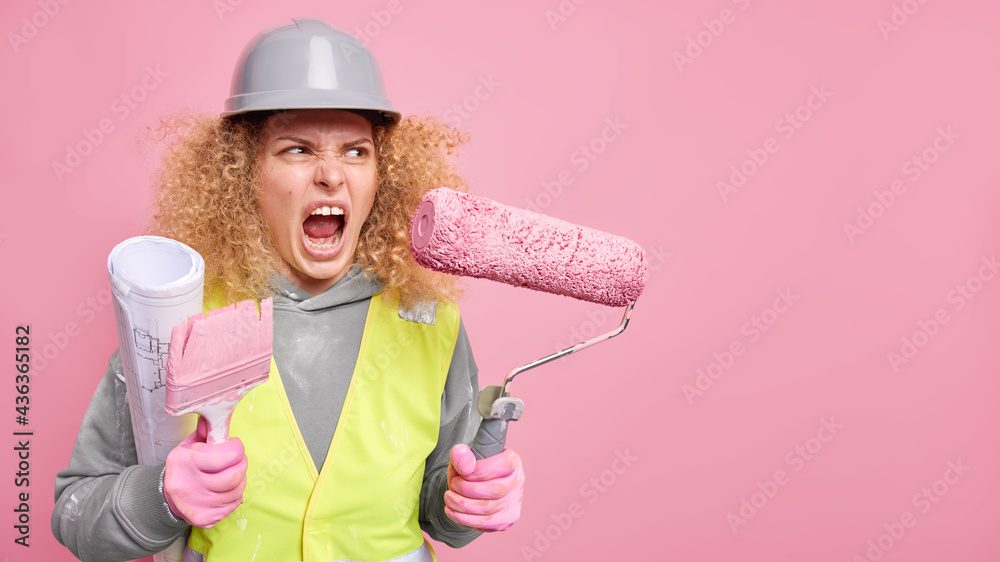 Angry irritated female construction worker holds building equipment and blueprint wears protective hardhat and safety clothes isolated over pink background with copy space. Occupation maitenance Angry irritated female construction worker holds building equipment and blueprint wears protective hardhat and safety clothes isolated over pink background with copy space. Occupation maitenance