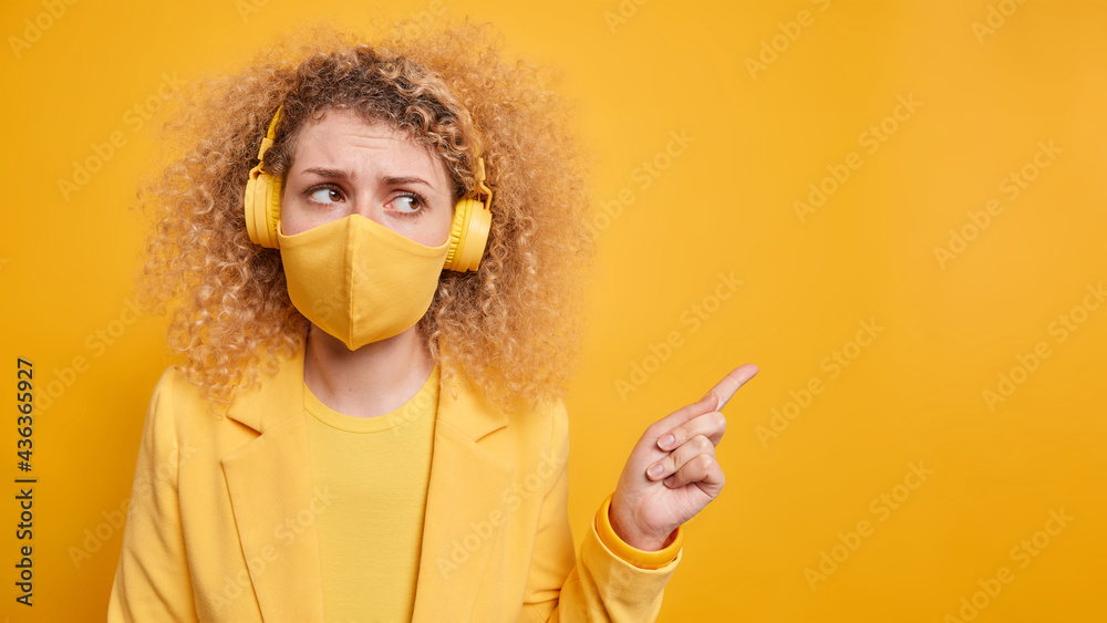 Photo of displeased young woman with curly hair wears protective face mask indicates at blank space shows space for your advertisement wears wireless headphones isolated over yellow background Photo of displeased young woman with curly hair wears protective face mask indicates at blank space shows space for your advertisement wears wireless headphones isolated over yellow background