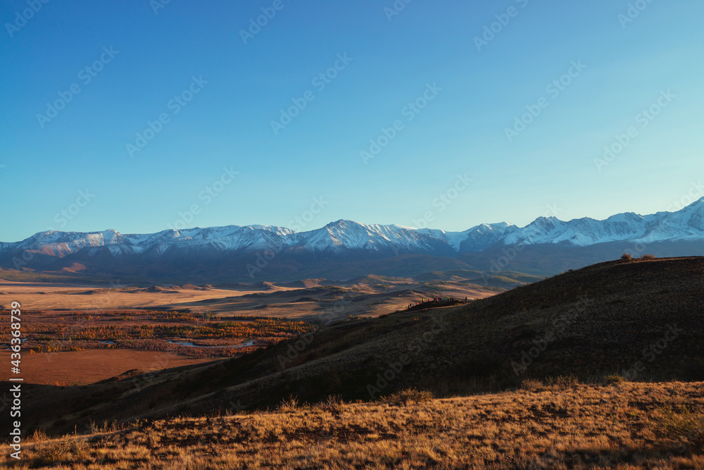 Awesome autumn landscape with tourists crowd on hill with view to red valley and great snow-covered mountain range in red sunset sunshine. Spectacular sunny view to snow mountains and autumn valley.
