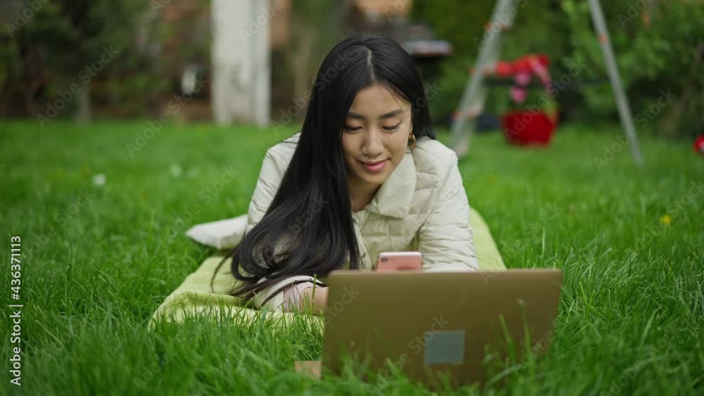 Front view of positive young Asian woman lying on green summer spring lawn with laptop texting on smartphone. Portrait of smiling beautiful slim millennial messaging online in garden outdoors Front view of positive young Asian woman lying on green summer spring lawn with laptop texting on smartphone. Portrait of smiling beautiful slim millennial messaging online in garden outdoors