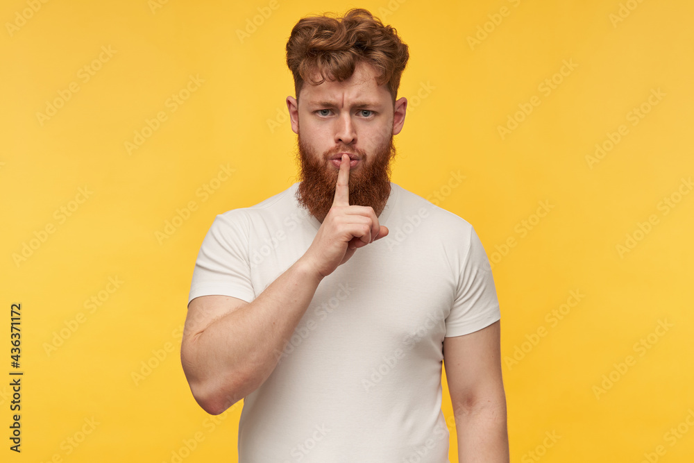 indoor portrait of young caucasian redhead man with a big heavy red beard, starring into camera, nervous and show silence gesture with a finger. Isolated over yellow bright background indoor portrait of young caucasian redhead man with a big heavy red beard, starring into camera, nervous and show silence gesture with a finger. Isolated over yellow bright background