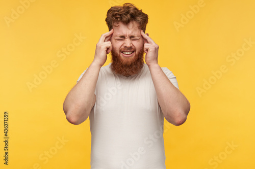 portrait of young redhead man with a big beard, keep hi fingers on temple, closed hi eyes, feels hard headache. isolated over yellow background