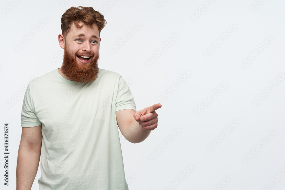 indoor shot of young man with a big beard and red hair, feels happy and smiling, while pointing with a finger at right side copy space. isolated over white background. indoor shot of young man with a big beard and red hair, feels happy and smiling, while pointing with a finger at right side copy space. isolated over white background.