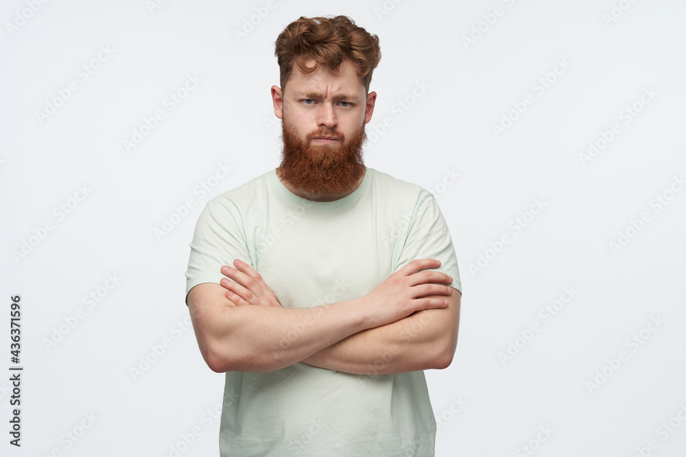 indoor shot of angry young redhead with a big beard male, crossed his hands on chest, looking into camera. pressed his lips and has negative facial expression.