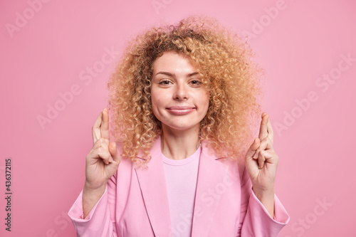 Young curly haired young woman crosses fingers believes in good luck smiles pleasantly pleads for dreams come true dressed in formal elegant clothes isolated over pink backround hopes or prays © wayhome.studio 