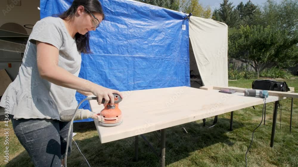 Woman sanding a large wooden tabletop with an electric sander. Woman uses sanding machine. Woman sanding a large wooden tabletop with an electric sander. Woman uses sanding machine.
