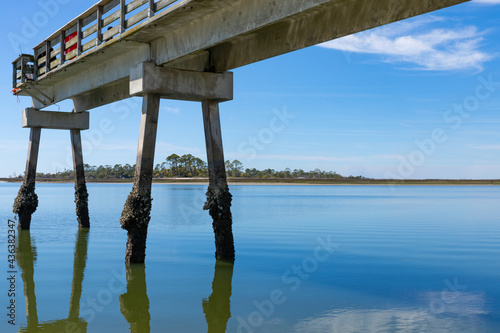 Narrow concrete fishing pier with oyster and barnacle covered pilings  blue sky reflected in still waters  Tybee Island Georgia USA  horizontal aspect