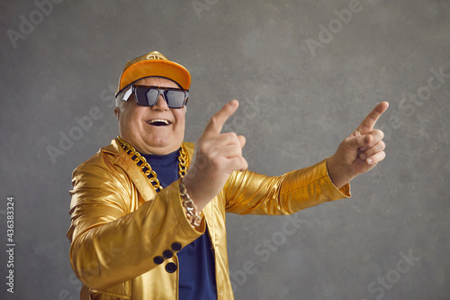 Funny happy rich senior man in golden party jacket, baseball cap and bling gold chain necklace enjoying rap music. Old but youthful grandpa dancing and having fun isolated on gray studio background