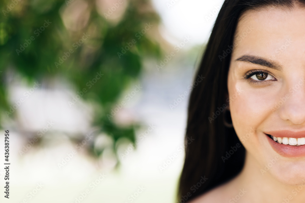 Close up half face portrait smiling attractive woman model looking at camera, happy beautiful 20s brunette lady pretty face dental smile posing stand alone at park on a background of sakura trees Close up half face portrait smiling attractive woman model looking at camera, happy beautiful 20s brunette lady pretty face dental smile posing stand alone at park on a background of sakura trees