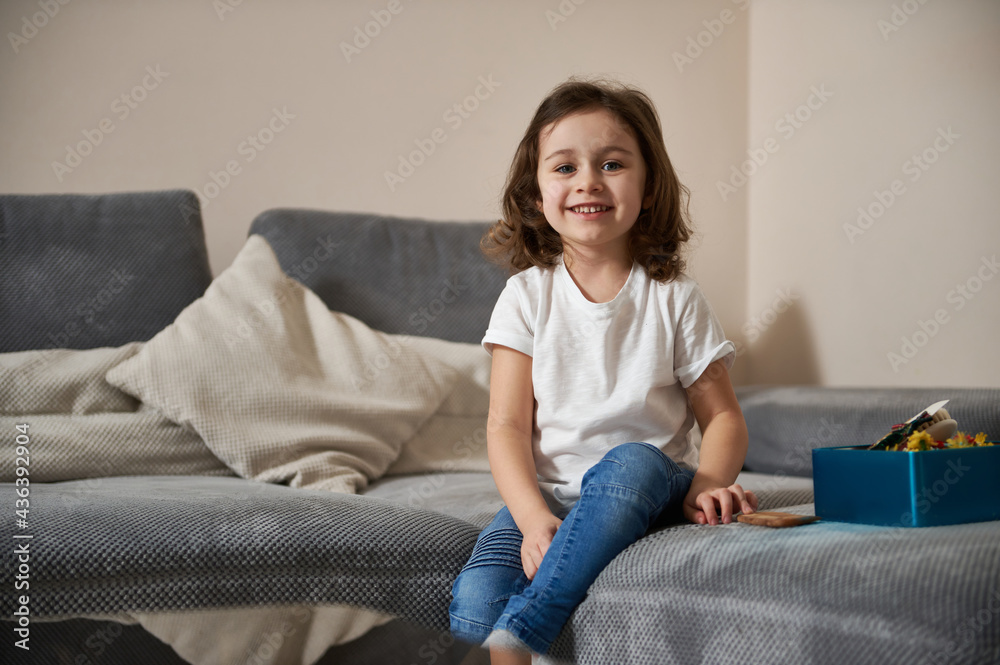Portrait of adorable preschool girl in casual clothes sitting on a sofa in the living room