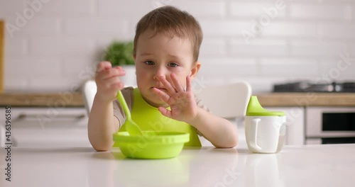 Cute baby boy does not want to eat, waves his head. Little child sitting on chair at home kitchen.