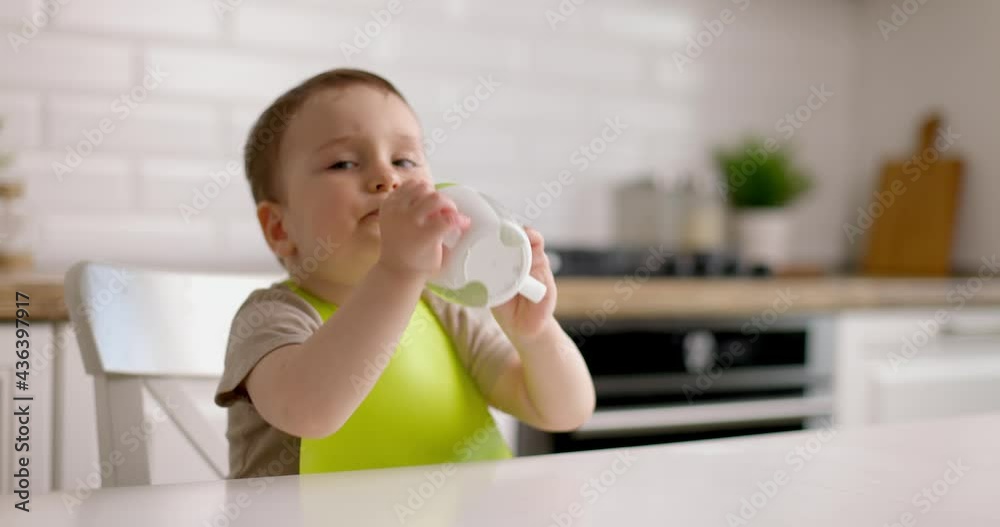 Cute baby boy sits at a table in the kitchen drinking from a kids' mug. Cute baby boy sits at a table in the kitchen drinking from a kids' mug.