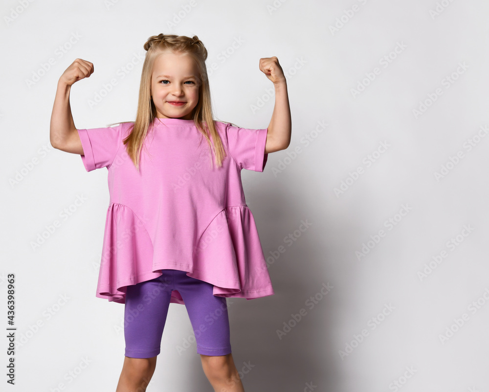 Funny cute little girl shows tense muscles on a white background. Super child in stylish summer clothes shows that she is ready for school or kindergarten. Place for text.