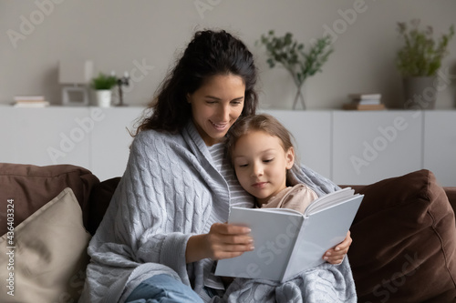 Loving young Latino mother and small biracial daughter sit relax on sofa in living room read book together. Caring happy Hispanic mom and little girl child rest at home on weekend enjoy literature.