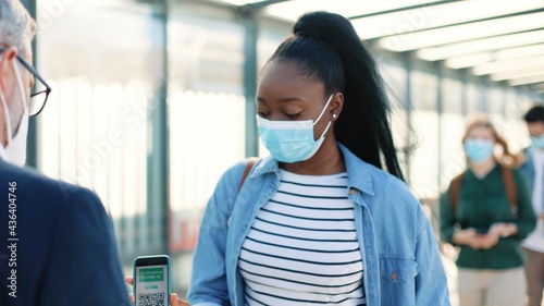 Close up of Caucasian man controller in mask on street checking health control scanning QR code of vaccination digital passport on smartphone app and measuring temperature of afro tourist © VAKSMANV