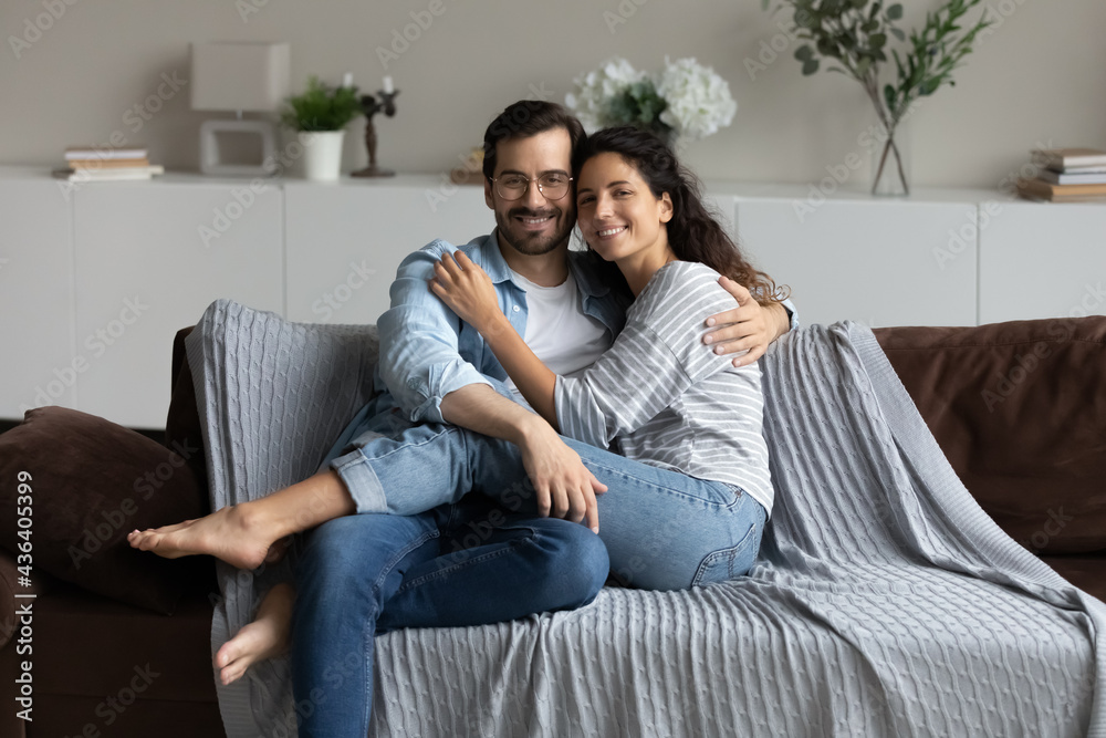 Portrait of happy multiracial man and woman renters sit relax on sofa in living room hugging. Smiling young multiethnic couple rest on couch at home enjoy relocation to new home. Rent concept.