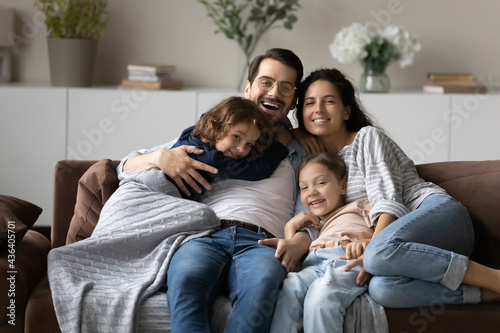 Portrait of overjoyed young multiethnic family with two small biracial children rest on sofa at home hugging cuddling. Happy multiethnic parents with little kids relax on couch. Rental, rent concept.
