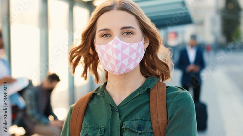 Camera approaching young pretty Caucasian positive woman standing with passport in city looking at camera at bus station. Close up of pretty female in mask travelling, coronavirus, covid pandemic