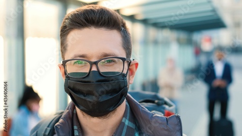 Camera approaching young Caucasian handsome happy man standing with backpack in city looking at camera at bus station with many people. Close up of guy traveller in mask outdoor, coronavirus pandemic