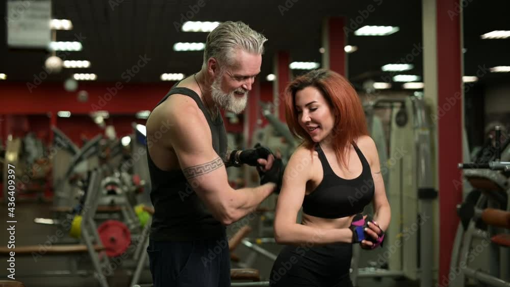 A gray-haired bearded man with tattoos poses together with a young athletic woman on the background of the gym. Satisfied athletes Show muscles to each other and rejoice at success A gray-haired bearded man with tattoos poses together with a young athletic woman on the background of the gym. Satisfied athletes Show muscles to each other and rejoice at success