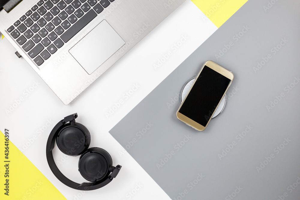 Yellow headphones, smartphone, gray laptop and wireless charger on trendy yellow gray background. Music, technology concept. Yellow headphones, smartphone, gray laptop and wireless charger on trendy yellow gray background. Music, technology concept.