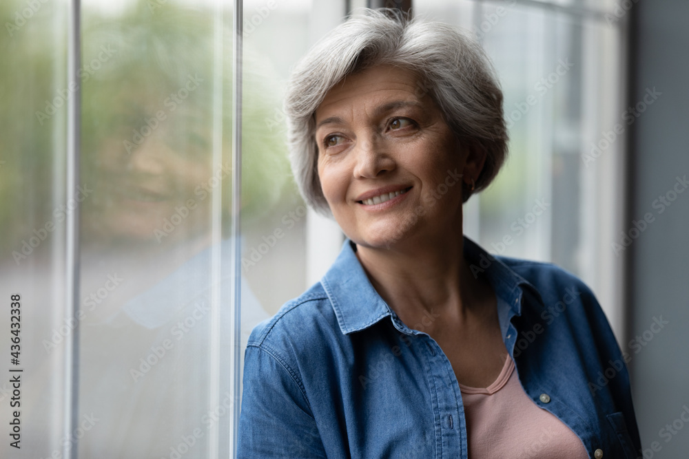Attractive smiling middle aged woman standing at window and looking outside. Grey-haired serene calm grandmother daydreaming pose alone indoor. Older generation good-looking person portrait concept Attractive smiling middle aged woman standing at window and looking outside. Grey-haired serene calm grandmother daydreaming pose alone indoor. Older generation good-looking person portrait concept