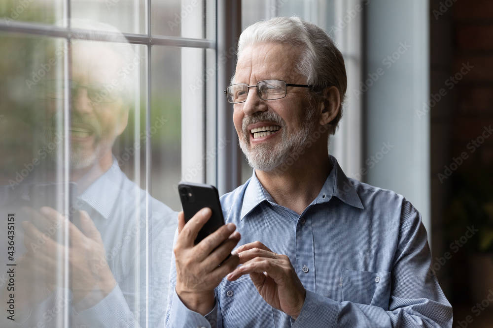 Cheerful older man enjoy modern tech usage standing near window looks outside feels happy. Hoary male in glasses holding smartphone making order, spend retired life with wireless technology concept Cheerful older man enjoy modern tech usage standing near window looks outside feels happy. Hoary male in glasses holding smartphone making order, spend retired life with wireless technology concept