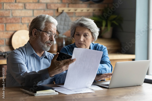 Tapety artystyczne Focused worried grey haired elderly spouses sit in kitchen with laptop on table reading financial papers documents, checking bills looking concerned, planning budget expenses, discussing debt concept