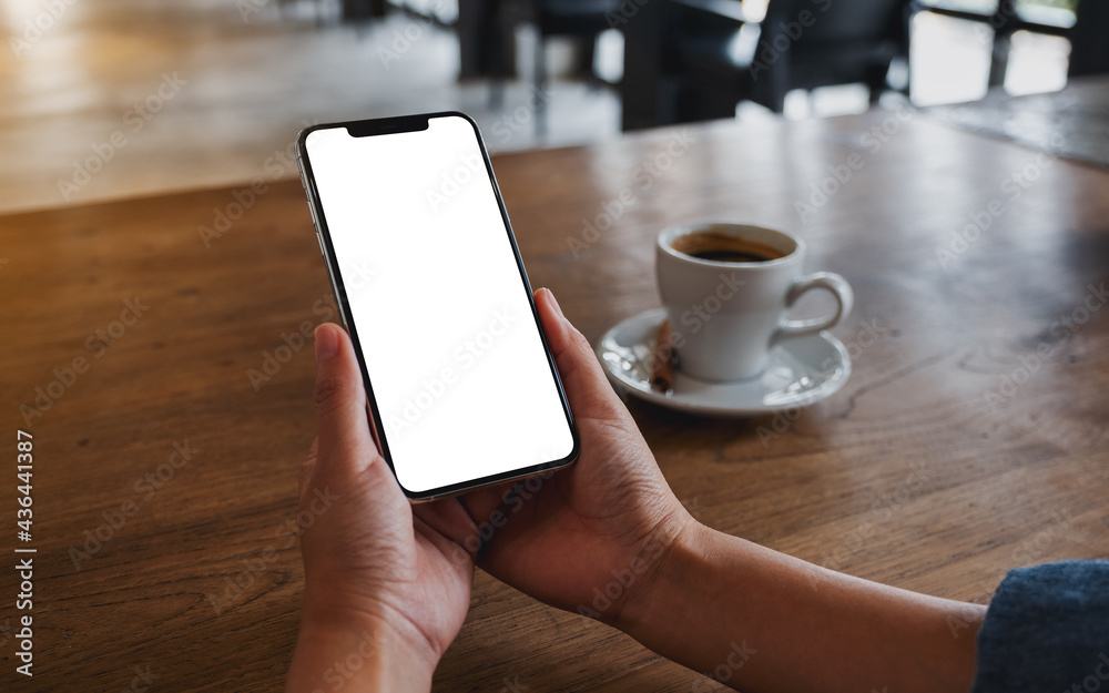 Mockup image of a woman holding mobile phone with blank white desktop screen with coffee cup on the table