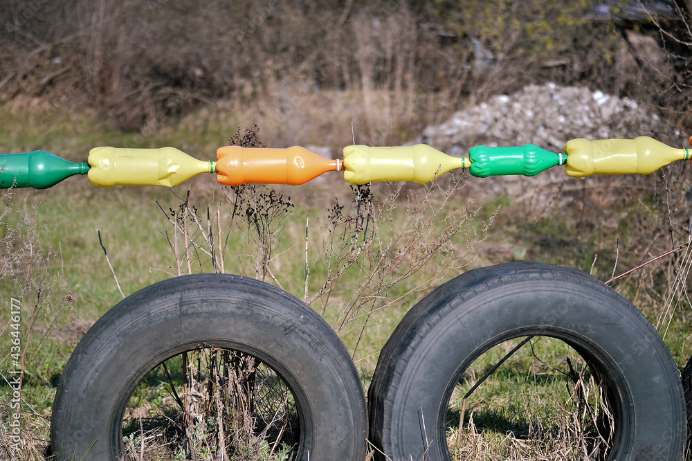 Fototapeta premium Plastic colored bottles hang in a row over black car tires against the backdrop of spring nature and piles of rubble in the background.