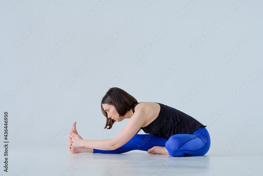 A young woman training yoga doing head to knee forward bend exercise, Janu Sirsasana pose in blue sports leggings and black top, on white background isolated on cyclorama. Healthy lifestyle.