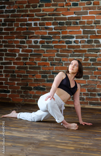 Beautiful young woman with white sporty pants and black top practicing hatha yoga Parivrtta Anjaneyasana pose crescent lunge on knee with twist, against the background of a brick wall in the loft. 