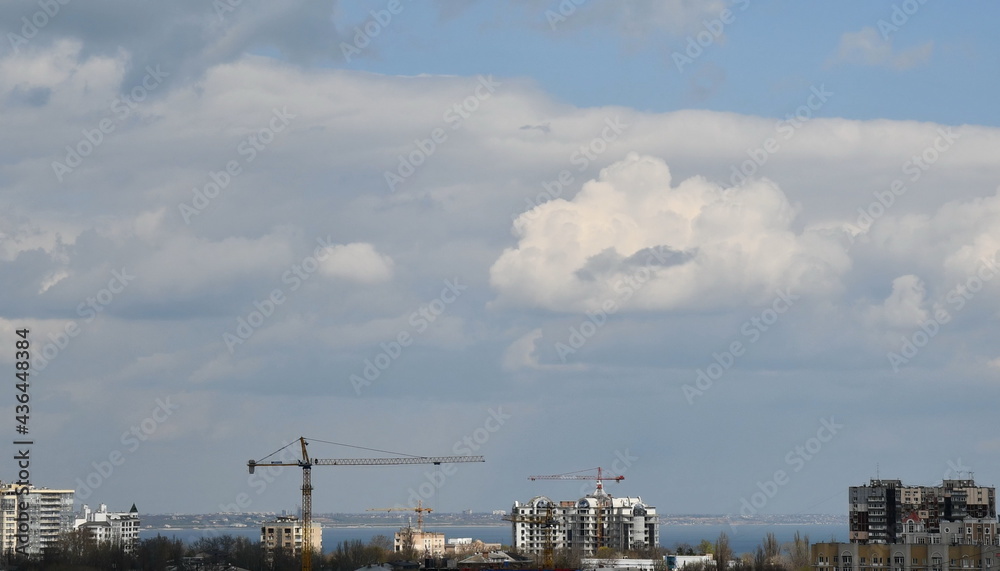 Construction crane jibs over rooftops under cloudscape. Under construction condo buildings in European city. Heavy construction machinery at urban skyline with cumulus clouds Construction crane jibs over rooftops under cloudscape. Under construction condo buildings in European city. Heavy construction machinery at urban skyline with cumulus clouds