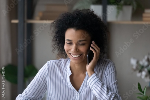 Happy millennial mixed race female laugh talk speak on cellphone gadget at home. Smiling young African American woman have fun engaged in pleasant smartphone conversation. Communication concept.